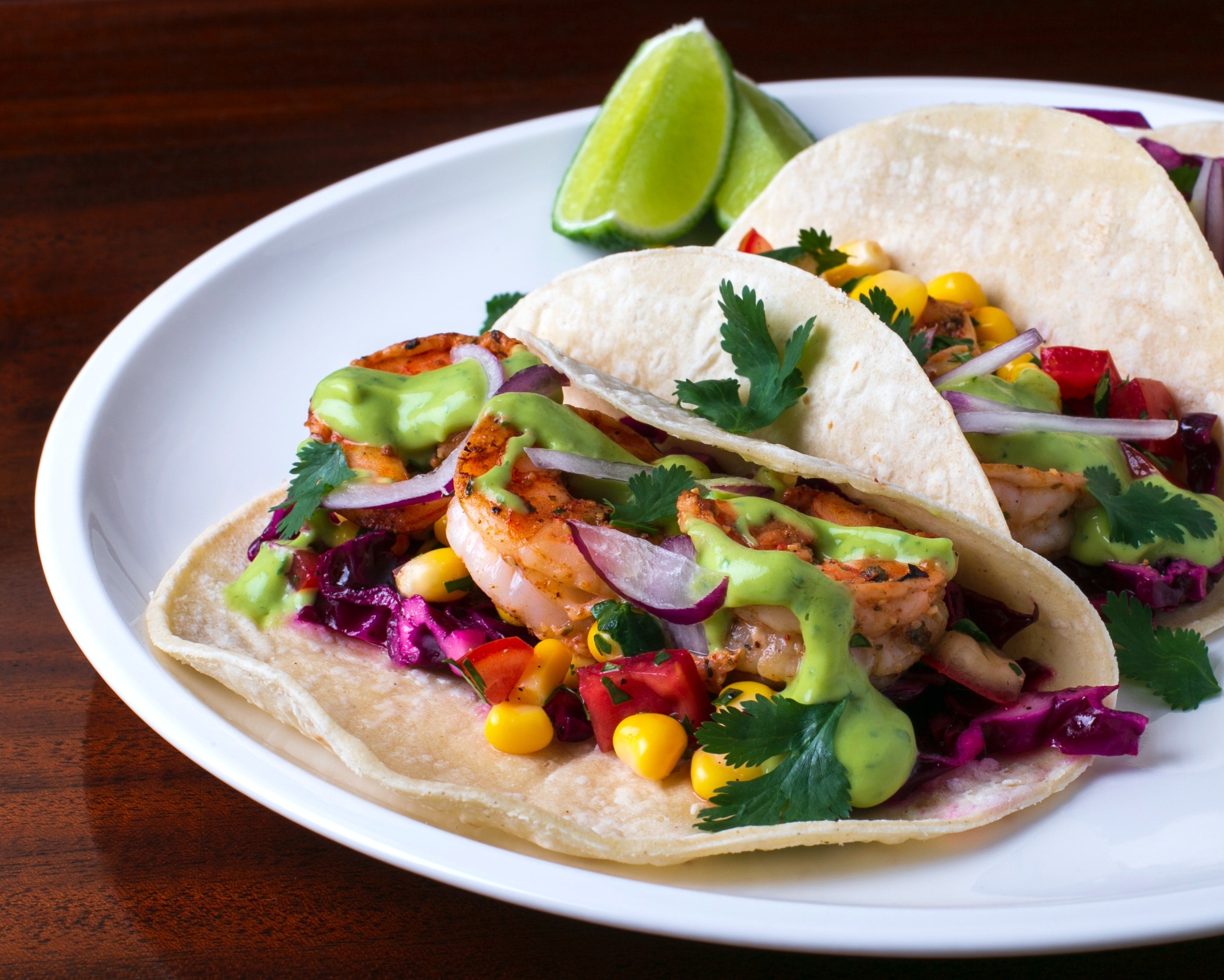 Shrimp tacos with colorful slaw, avocado crema and lime wedges on a white plate